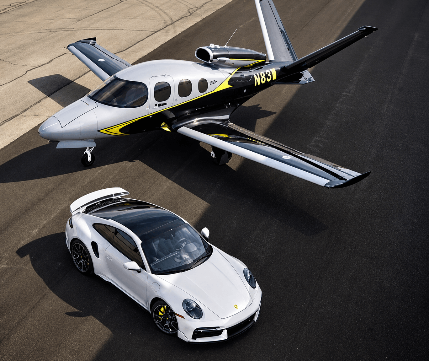 A white Porsche 911 parked beside a silver and black Cirrus Vision Jet SF50 on the ramp, seen from above.